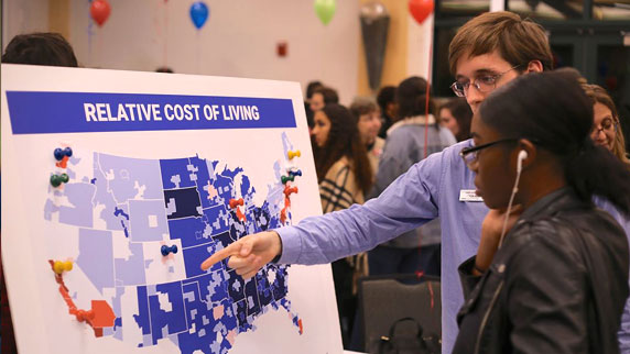 Two students standing in front of a board that shows a map of the United States and the relative cost of living for each state.