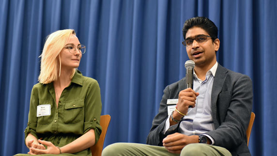 Two students sitting on a stage. One is talking into a microphone.