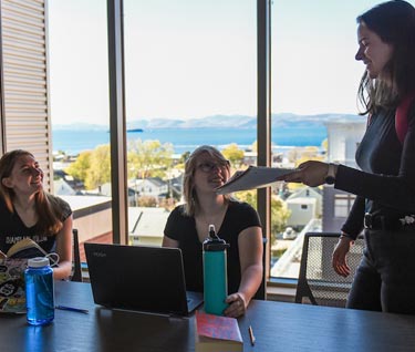Four students around a table in a common room with a view of Lake Champlain and mountains behind them