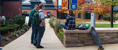 Students chatting on campus, two male students with backbacks one female student with a skateboard
