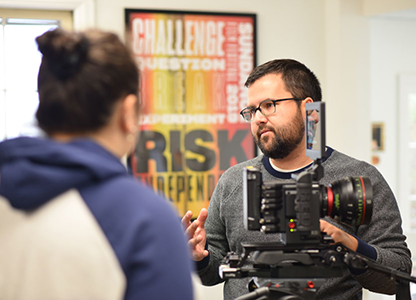Male student stands on one side of a professional video camera, speaking to another student 