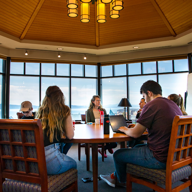 Students and a professor sit around a table in Champlain's library. View of Lake Champlain in the distance.