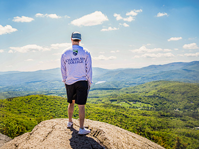 student in Champlain College t-shirt stand on a large rock overlooking a valley of trees