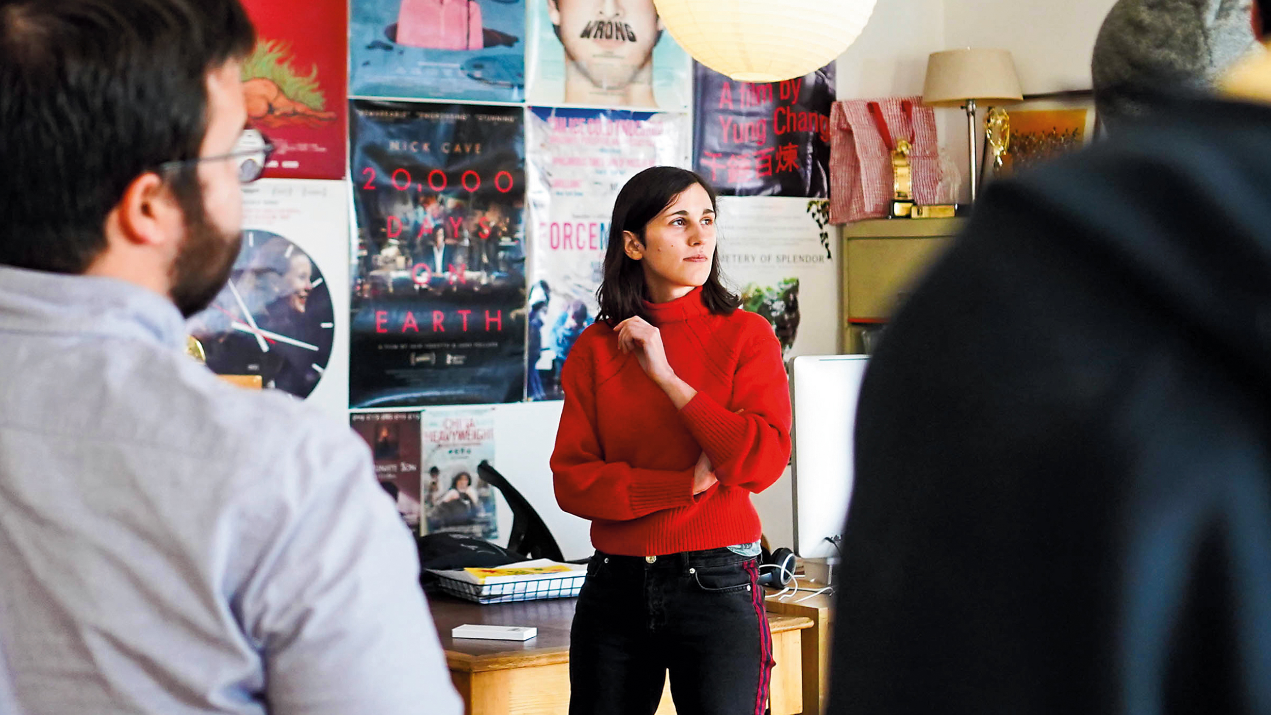 professor stands at the front of a group; wall full of posters in the background