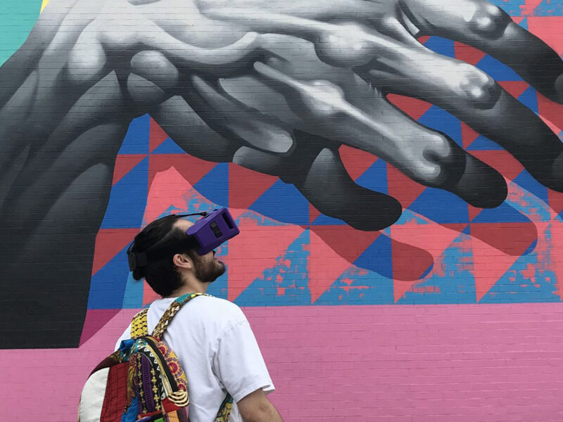 A student wearing what appears to be a VR headset looking towards a mural on a wall of a gray hand over a colorful background.