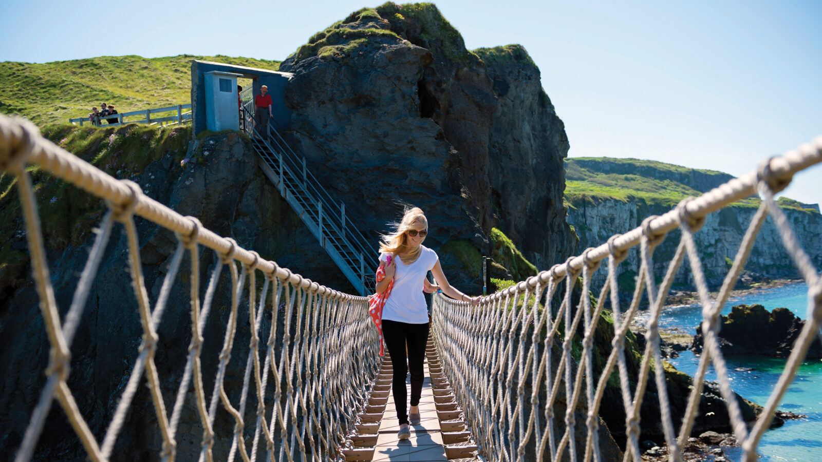 woman walks across rope bridge on cliffs near ocean in Ireland