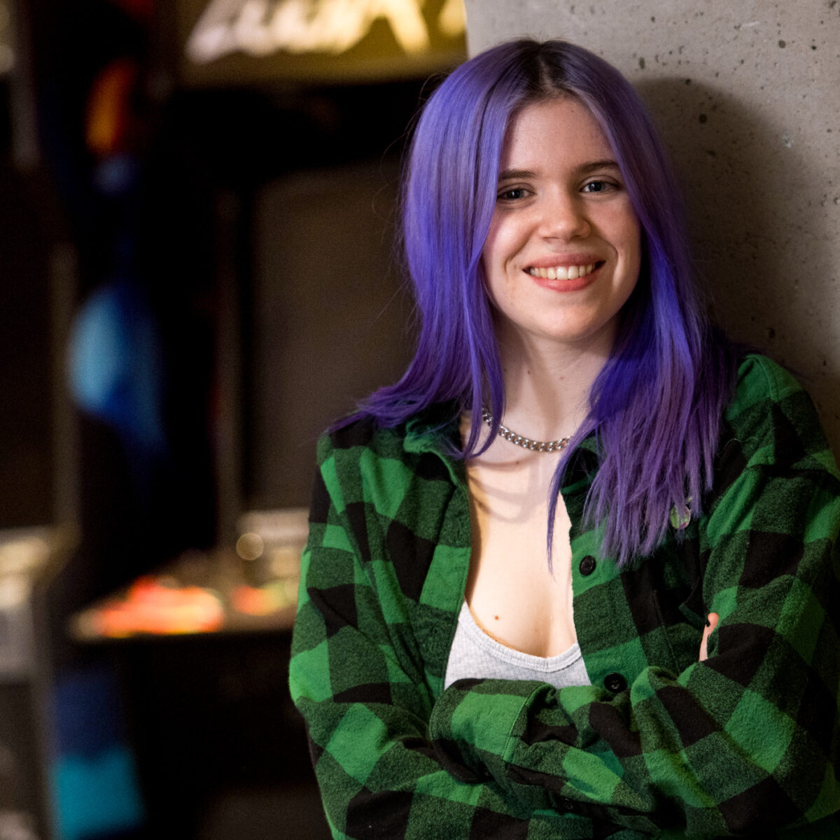 female student with purple hair stands in front of arcade games, smile at camera