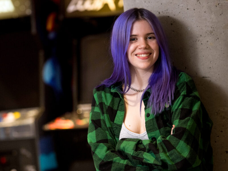 female student with purple hair stands in front of arcade games, smile at camera