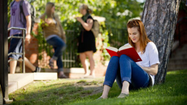student leans against a tree while reading a book
