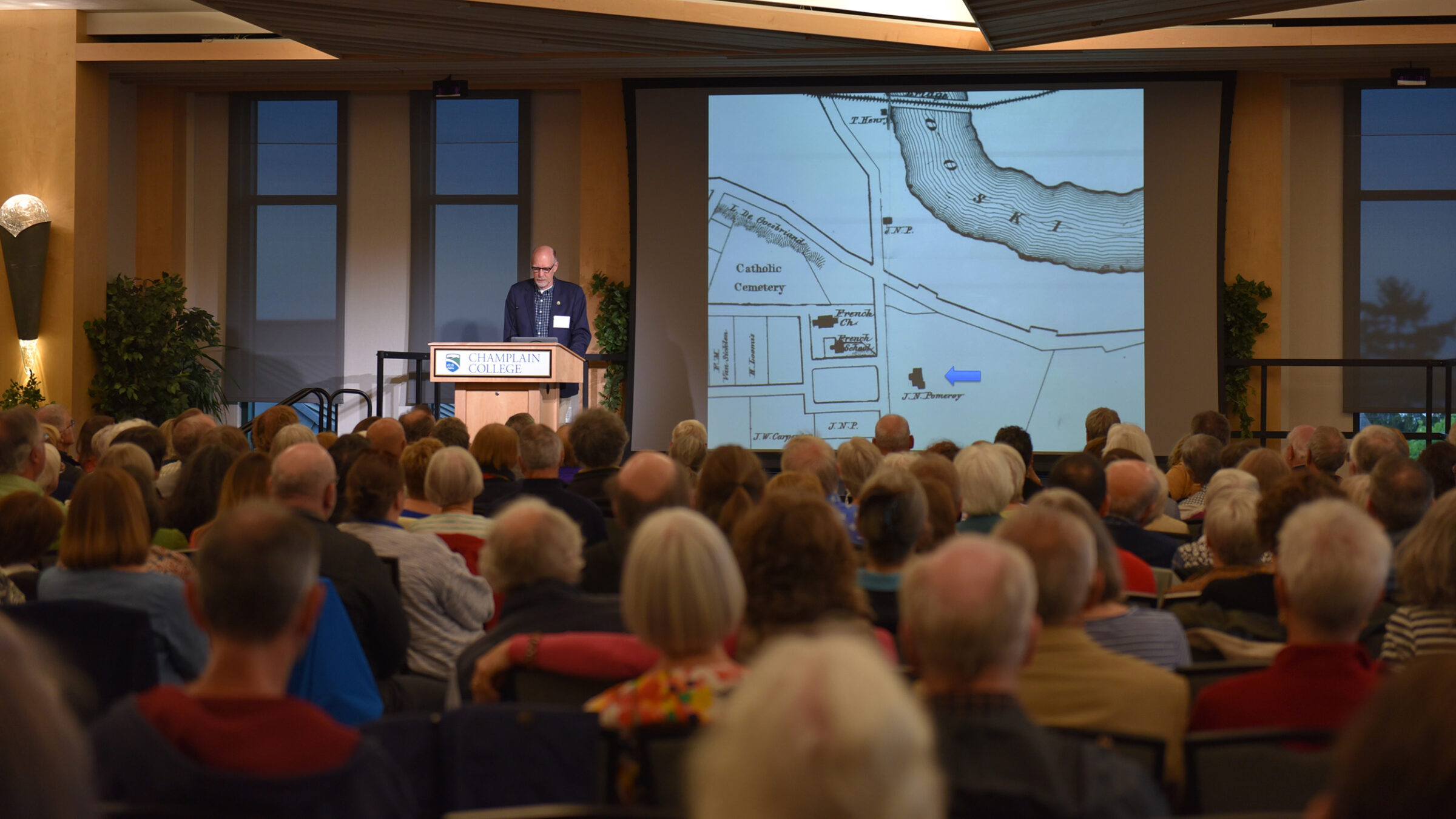 a speaker at a podium speaks to a group, a map on the screen