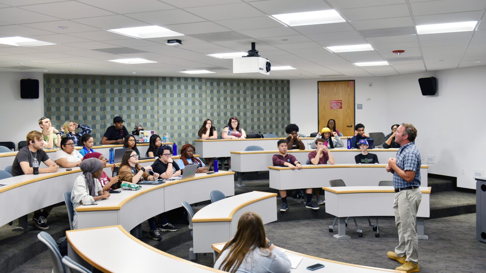A group of 25 students sit in a classroom with rounded tables listening to a professor speaking at the front of the room