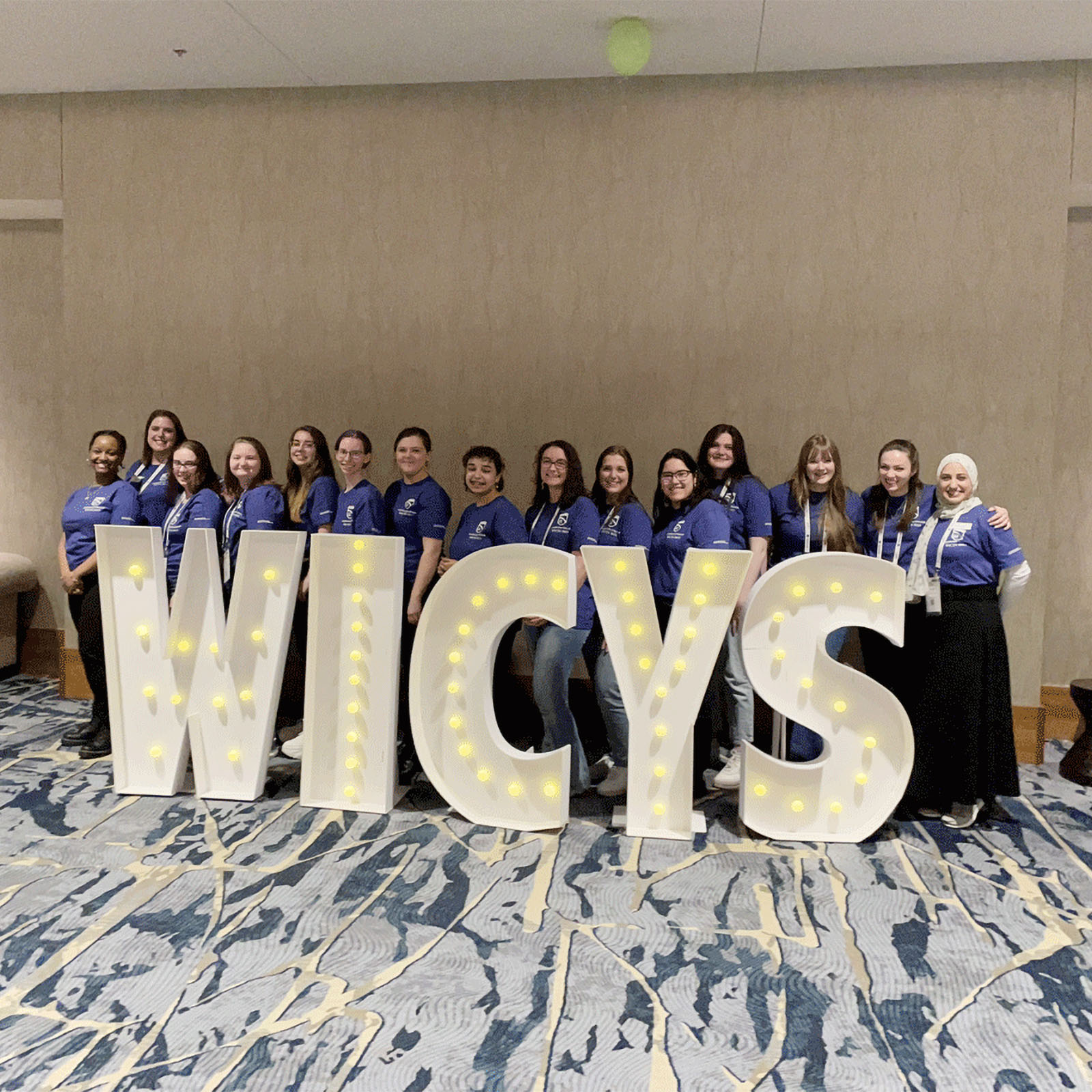 a large group of female cybersecurity students posing behind a "women in cybersecurity" sign at an event