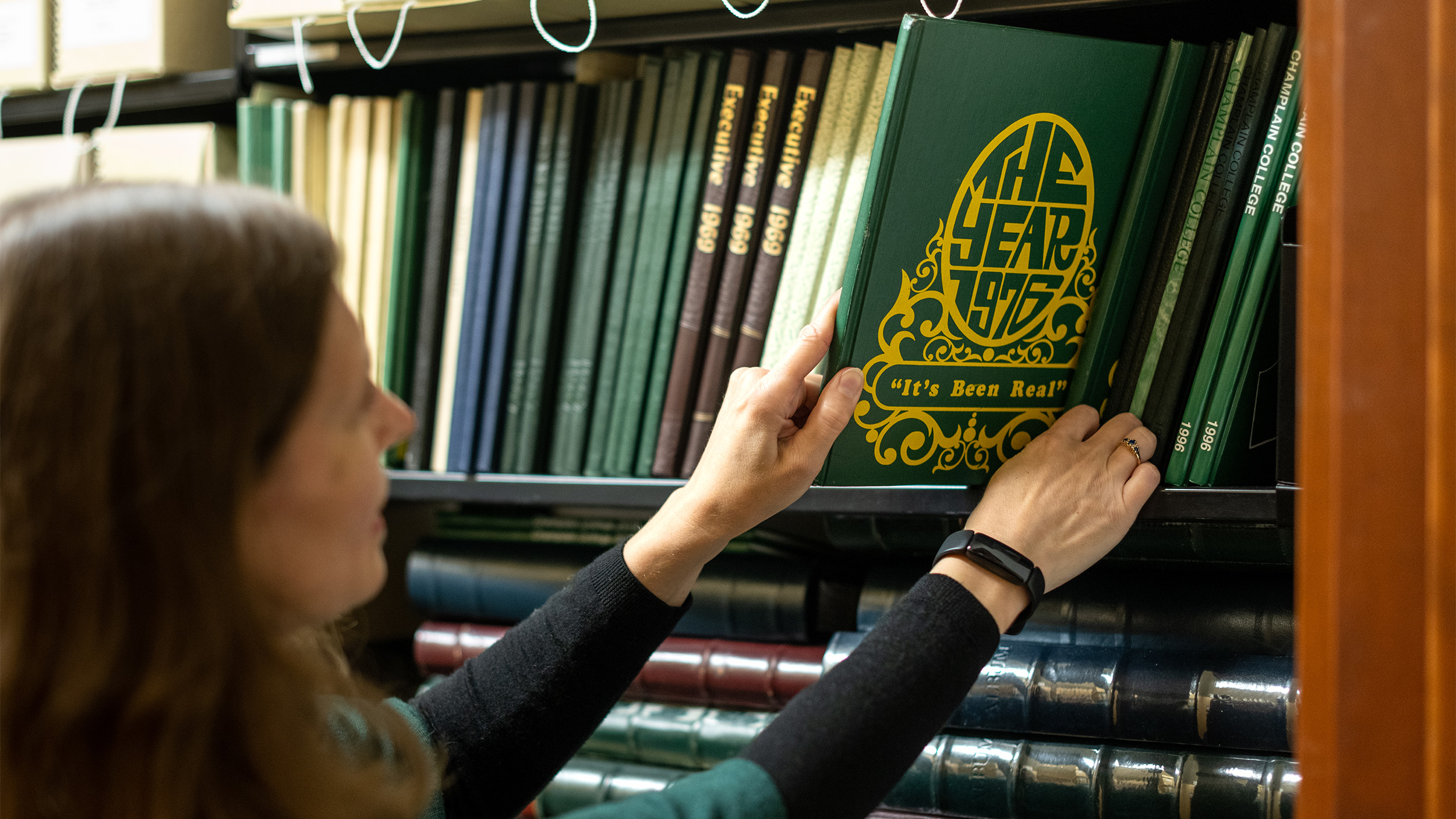 An employee removes the 1976 Champlain College yearbook from a shelf of yearbooks and other historic books