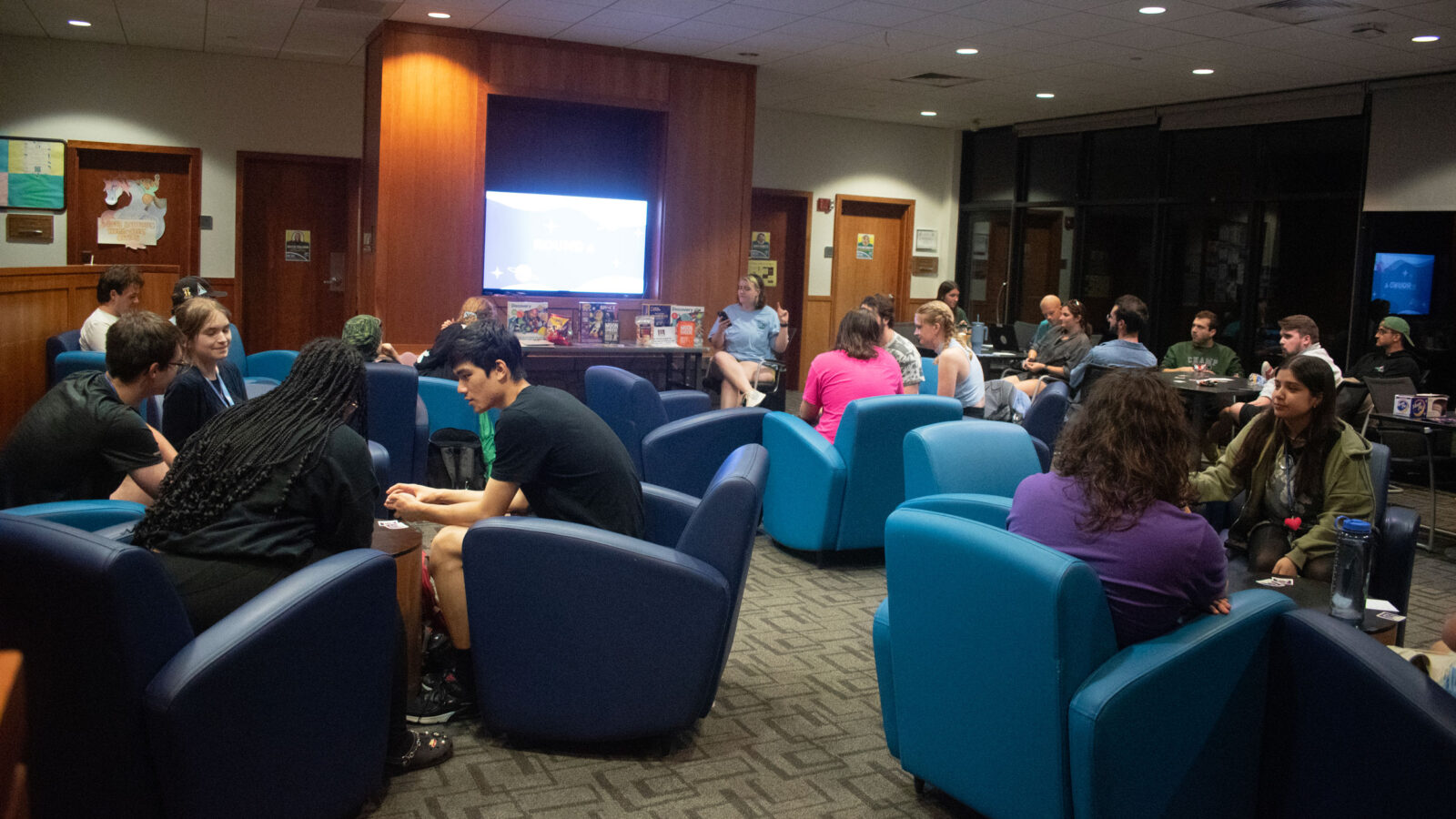 a group of about 20 students sit in blue chairs in fireside lounge playing a game in groups of 5