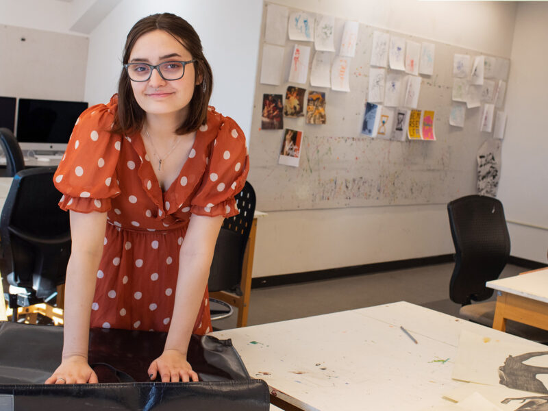 student stands over her art binder, looks at the camera