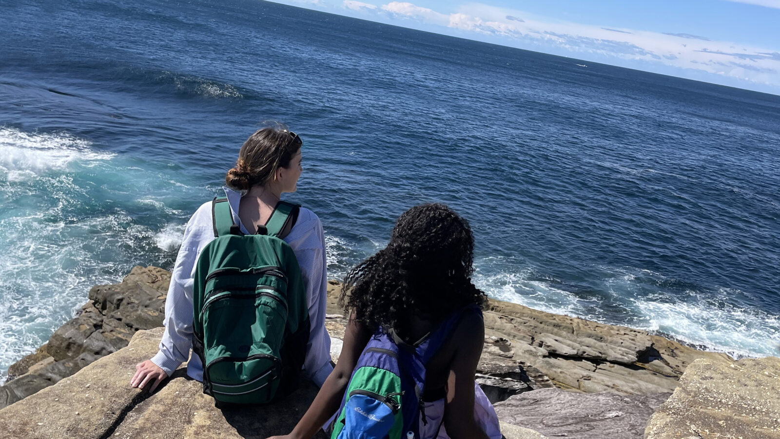two students sit on a rocky beach looking out at the ocean