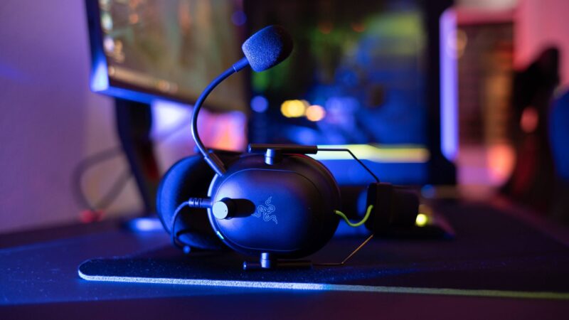 Razer headphones resting on a gaming desk with a pc and monitor lit by RGB lighting