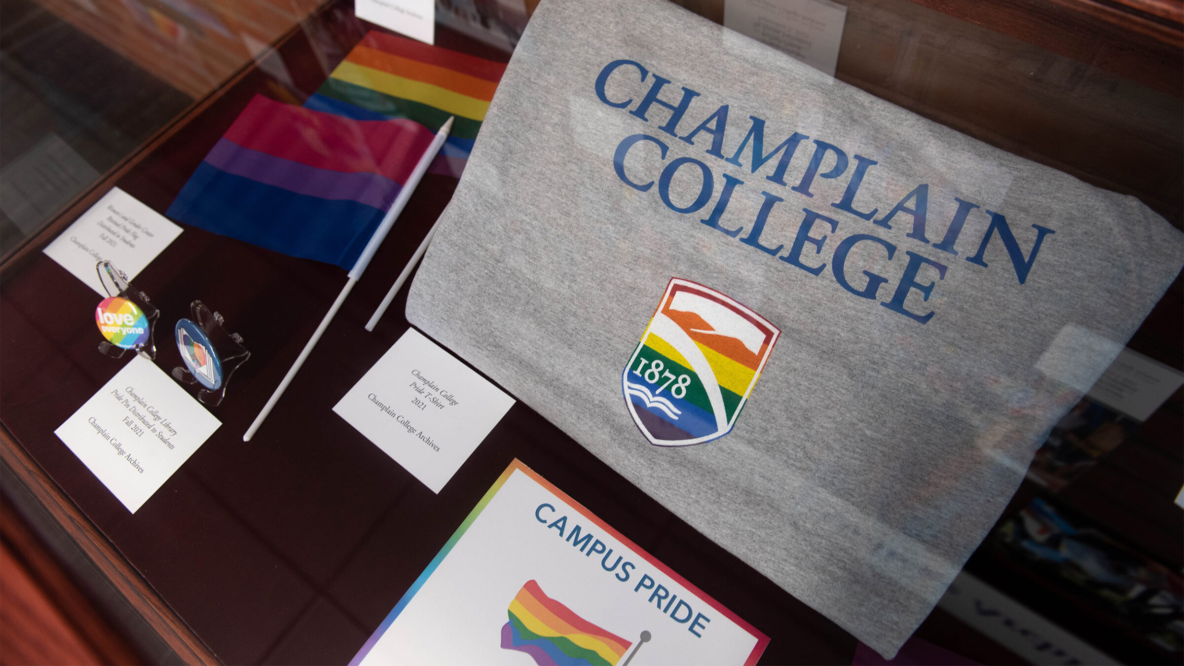 Part of a Campus Pride display including a Champlain College t-shirt with a rainbow-colored shield, two types of mini pride flags, and two pins.