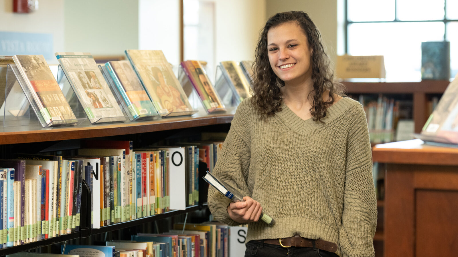 A student stands between two bookshelves in the library. She is holding a book and smiling. Other books are on display stands on the shelves.