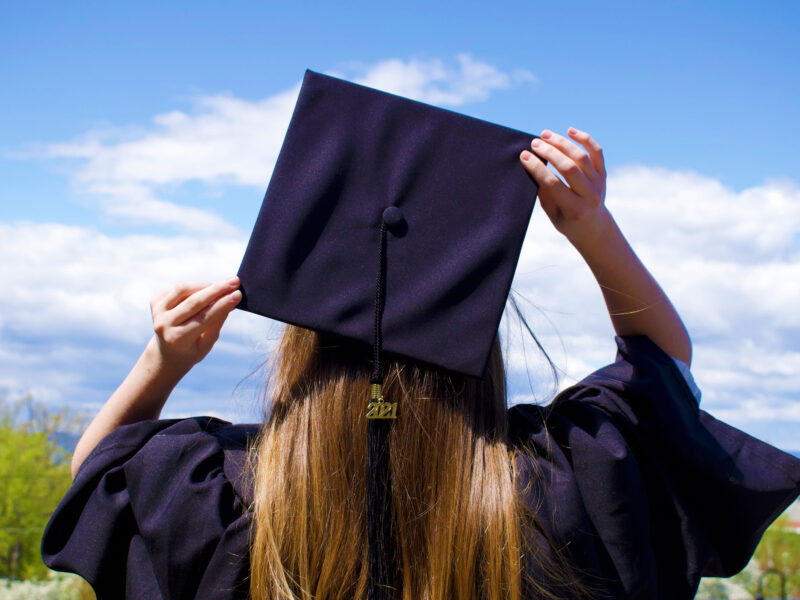 A student faces away from the camera in their graduation regalia, looking out at Lake Champlain.