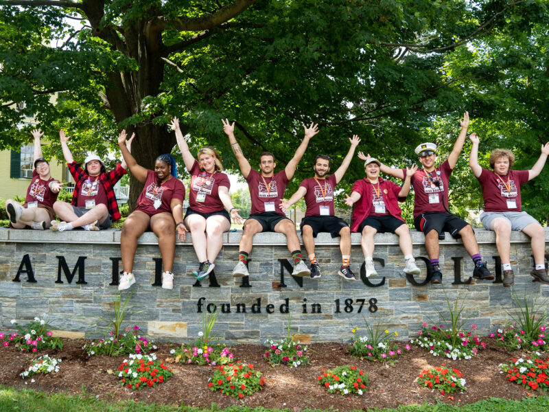 a group of student ambassadors sit on the champlain college sign outdoors wearing matching tshirts for orientation