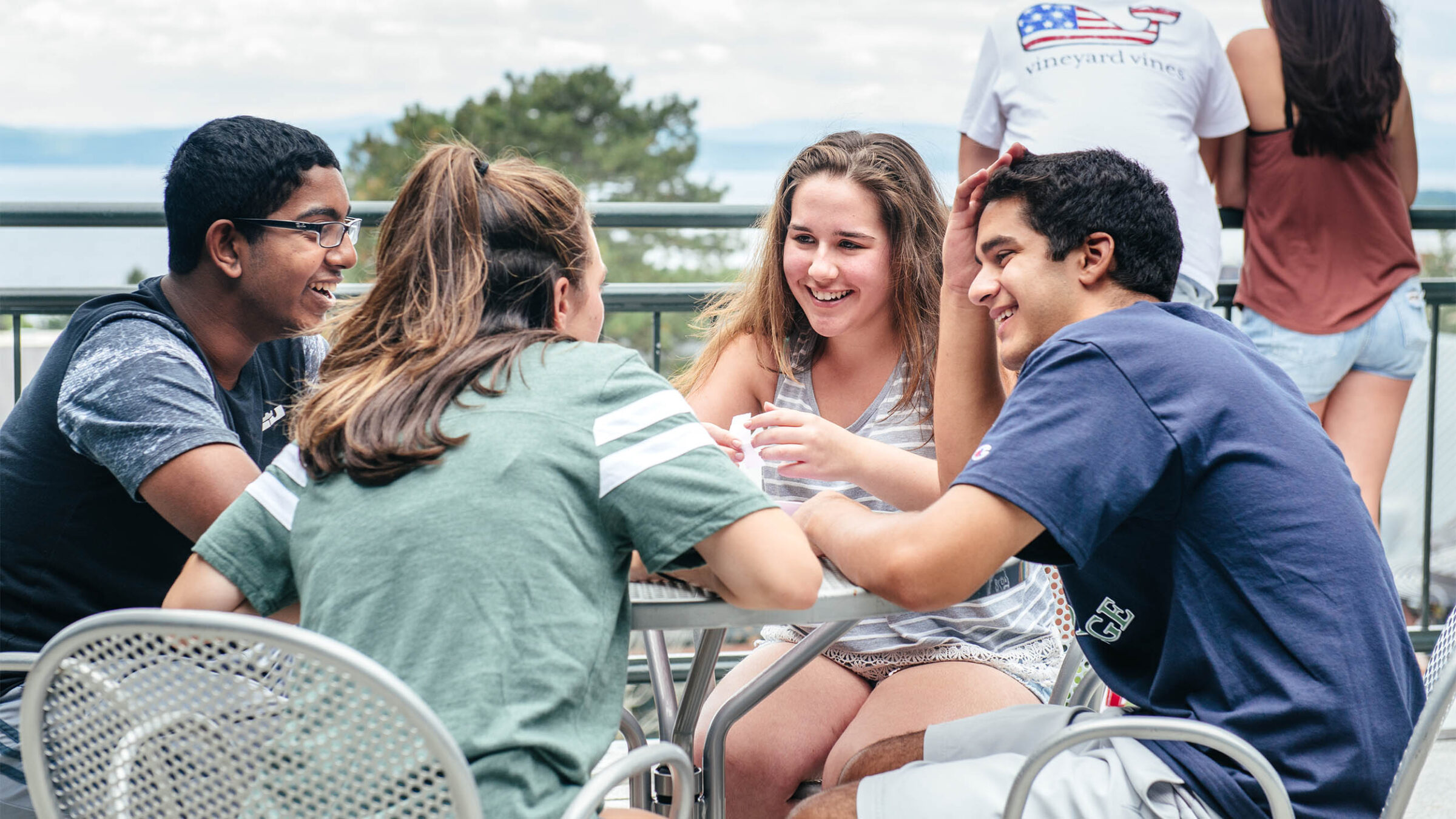 four smiling students sharing a conversation on an outdoor patio while two other students observe the view across the lake