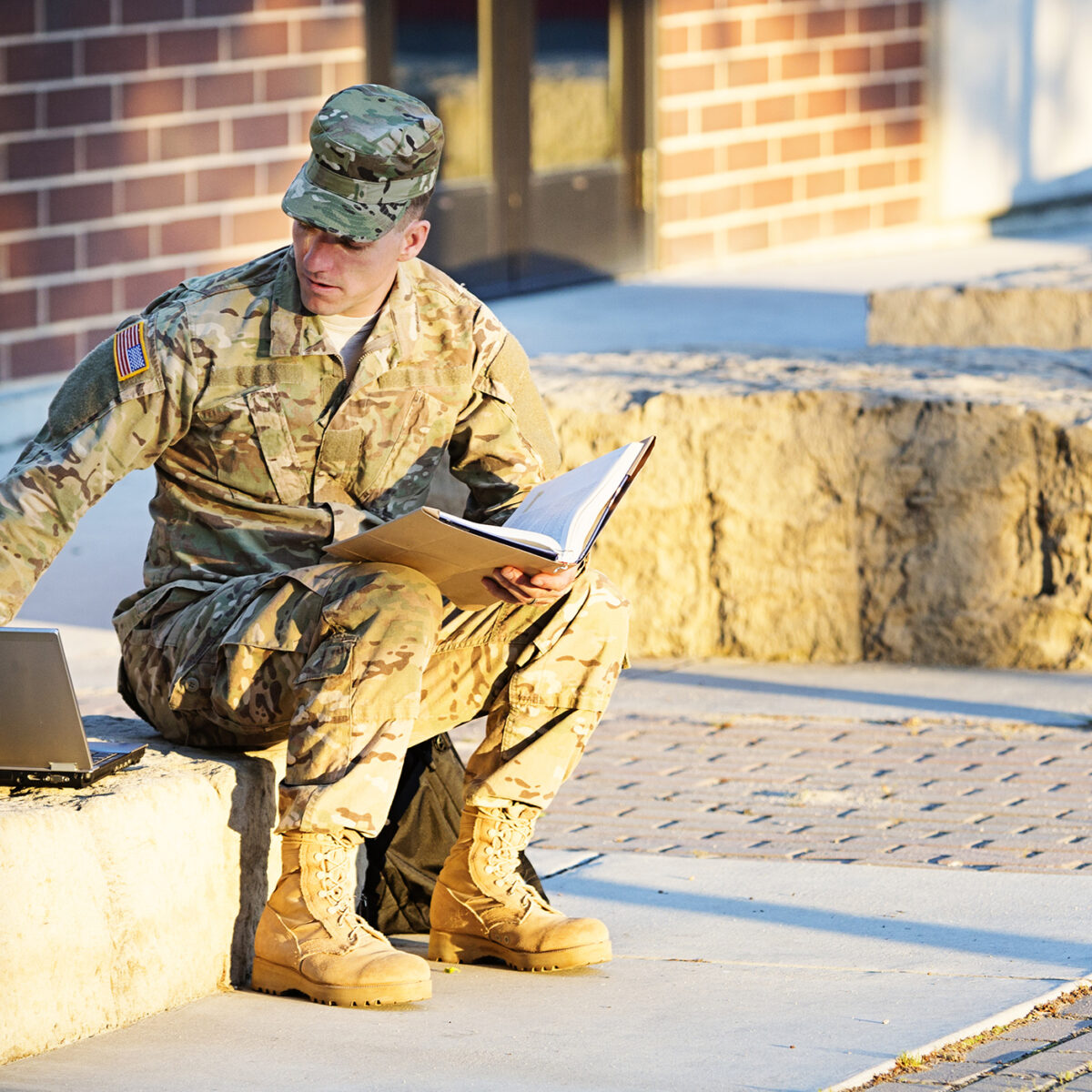 US soldier in camouflage uniform with laptop in front of campus