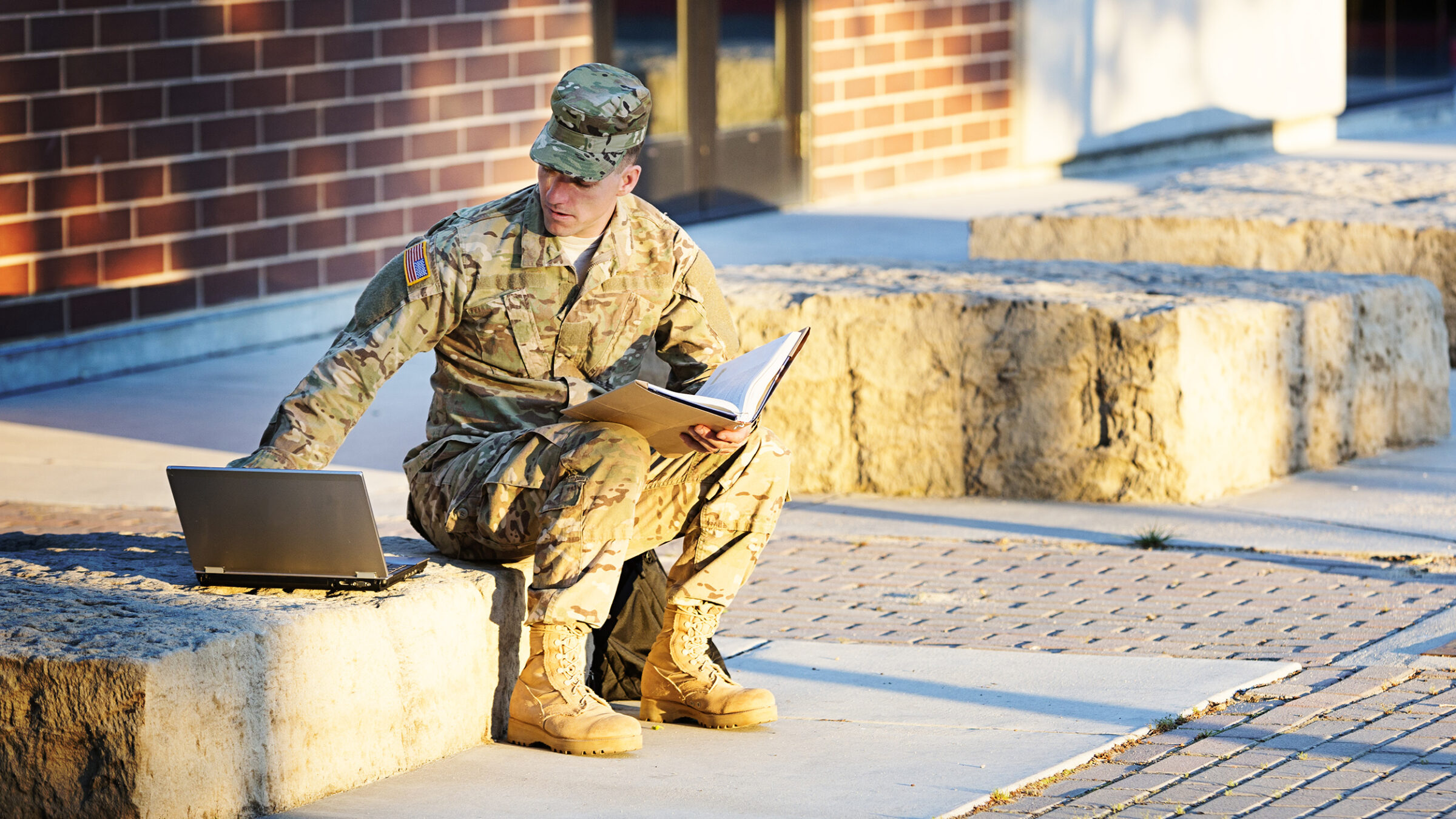 US soldier in camouflage uniform with laptop in front of campus