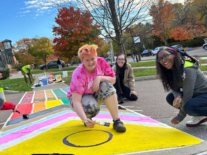 four students smile with paintbrushes in hand as they paint an intersex flag crosswalk on campus, with blue skies behind them