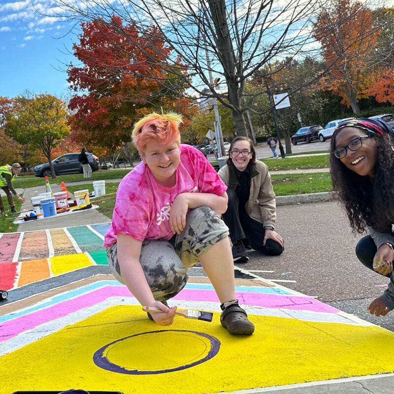 four students smile with paintbrushes in hand as they paint an intersex flag crosswalk on campus, with blue skies behind them