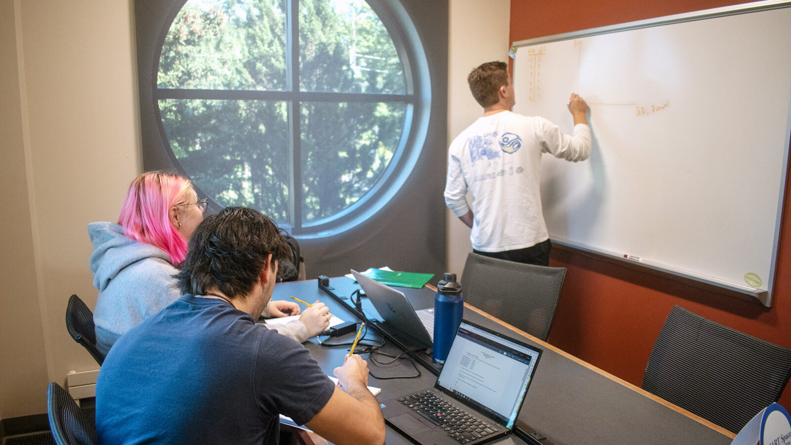 a student at a white board tutors 2 other students at a table