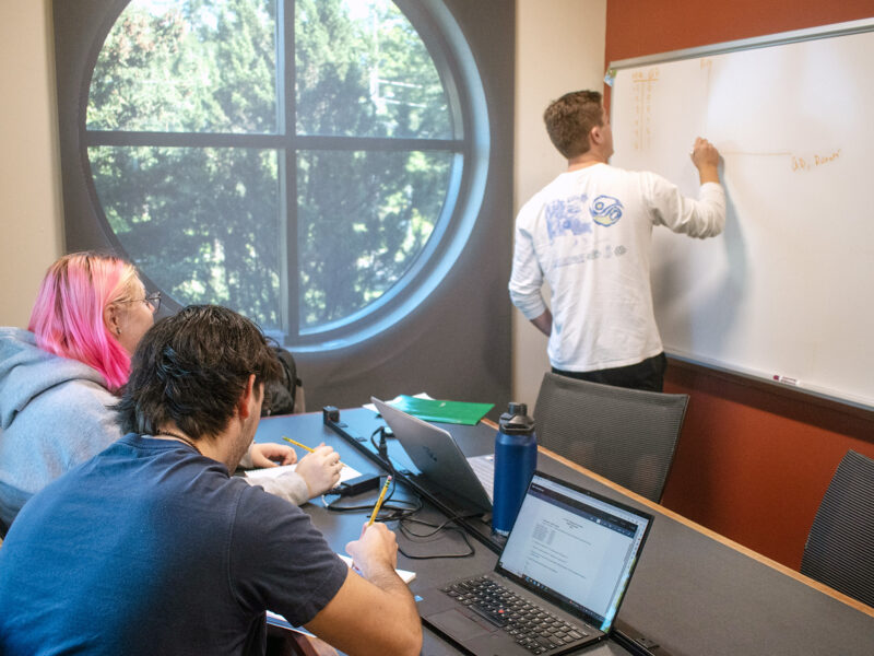 a student at a white board tutors 2 other students at a table