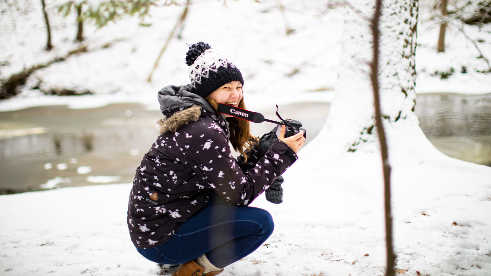 A student crouches in the snow to get a wintery photo in the woods.