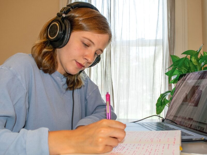 Student working at a desk with headphones on and a pen and notebook in hand in front of an open laptop