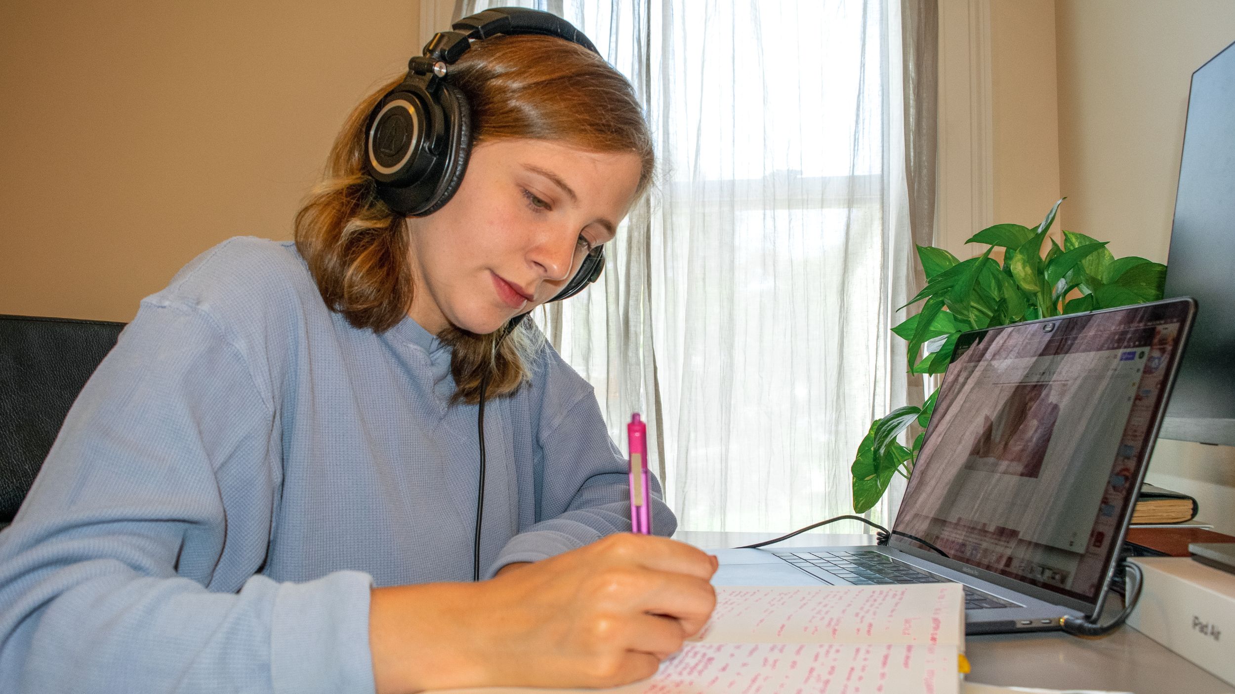 Student working at a desk with headphones on and a pen and notebook in hand in front of an open laptop
