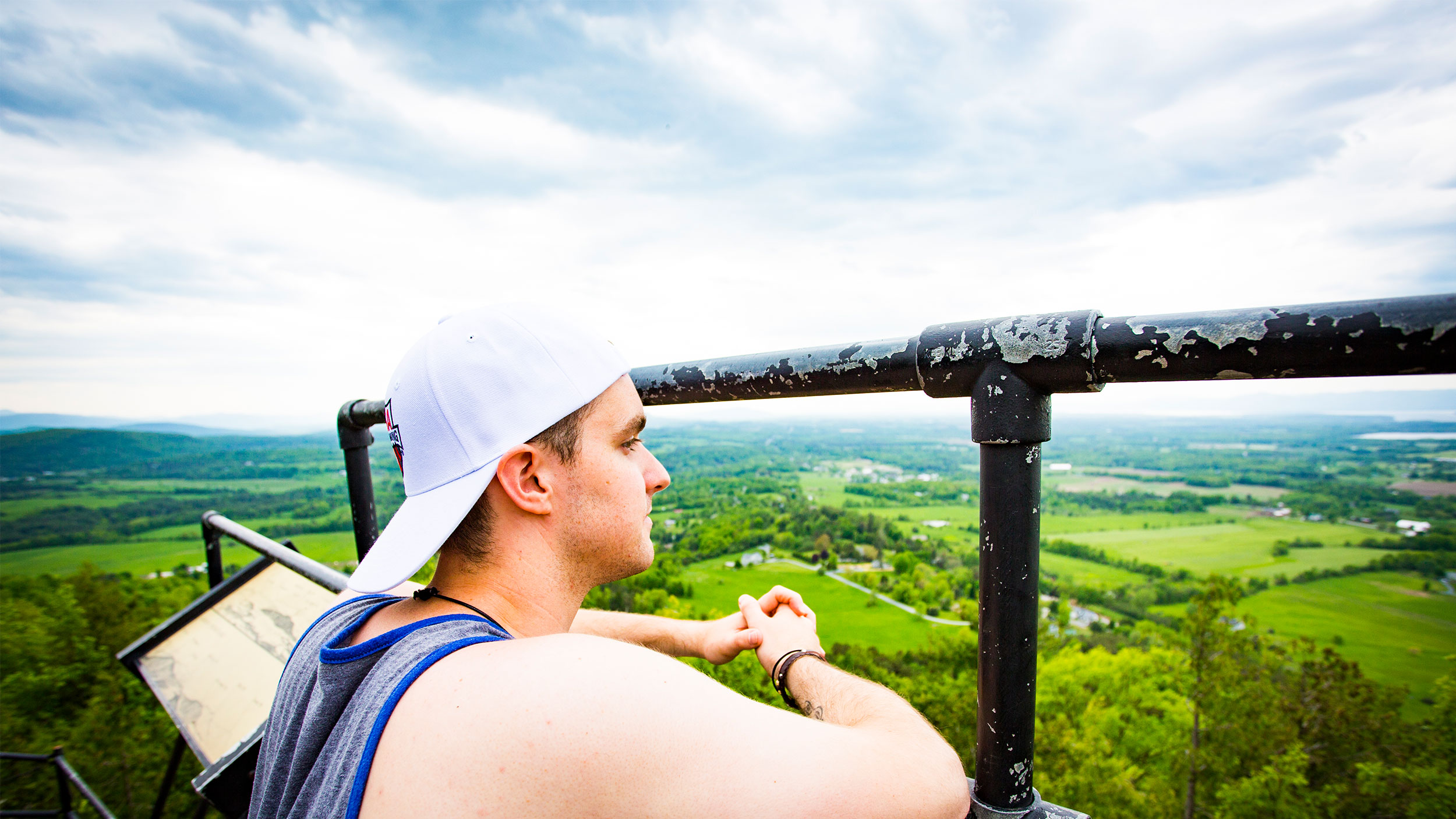 student wears a backward white baseball cap and stands at the top of mount philo behind a black fence, looking out on the green horizon