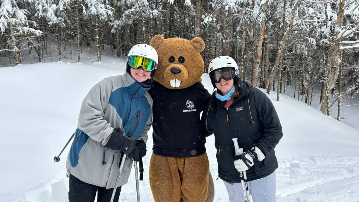 students and Chauncey posing for a photo on the ski mountain with helmets, goggles, and poles