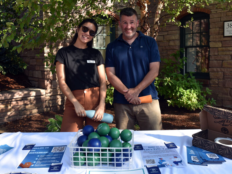 two champlain college counseling staff members stand and smile behind a table at the fall activities fair. a box of stress balls sit on the table covered in a white tablecloth.