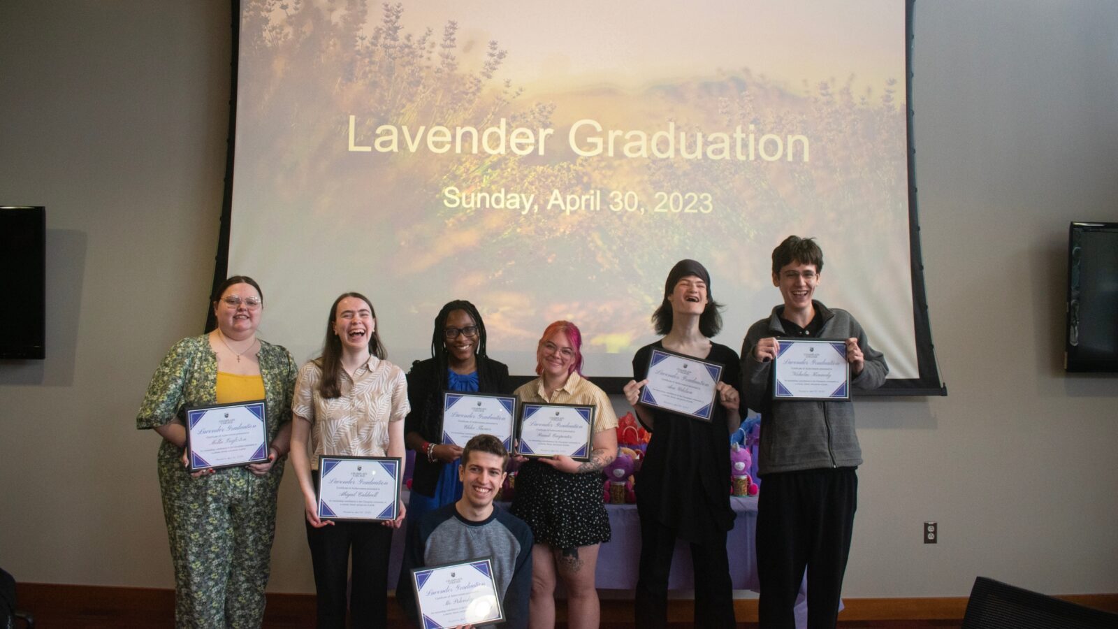 seven students hold framed certificates in front of a projector screen reading "lavender graduation sunday, april 30, 2023"