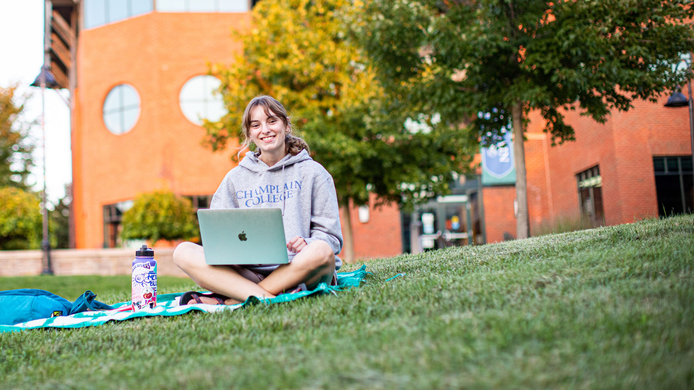 student sitting on the grass outside in a champlain college hooded sweatshirt with their laptop