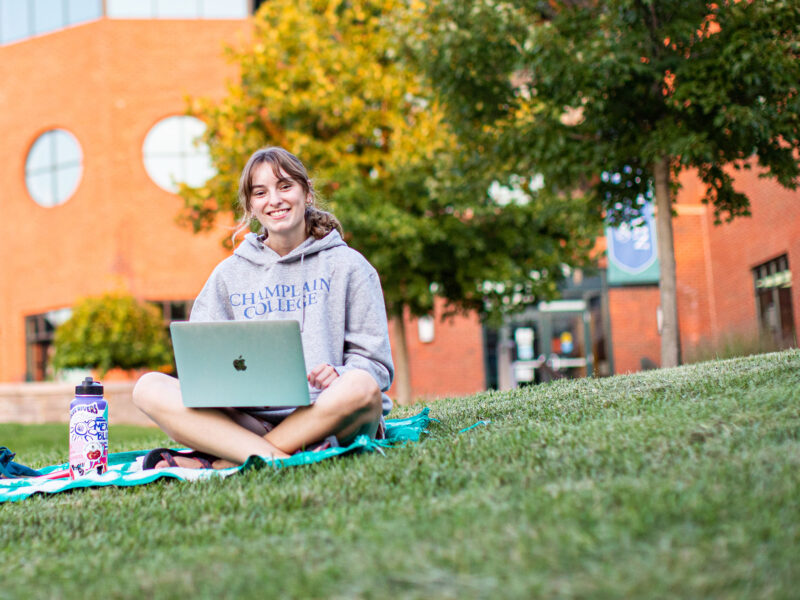 student sitting on the grass outside in a champlain college hooded sweatshirt with their laptop