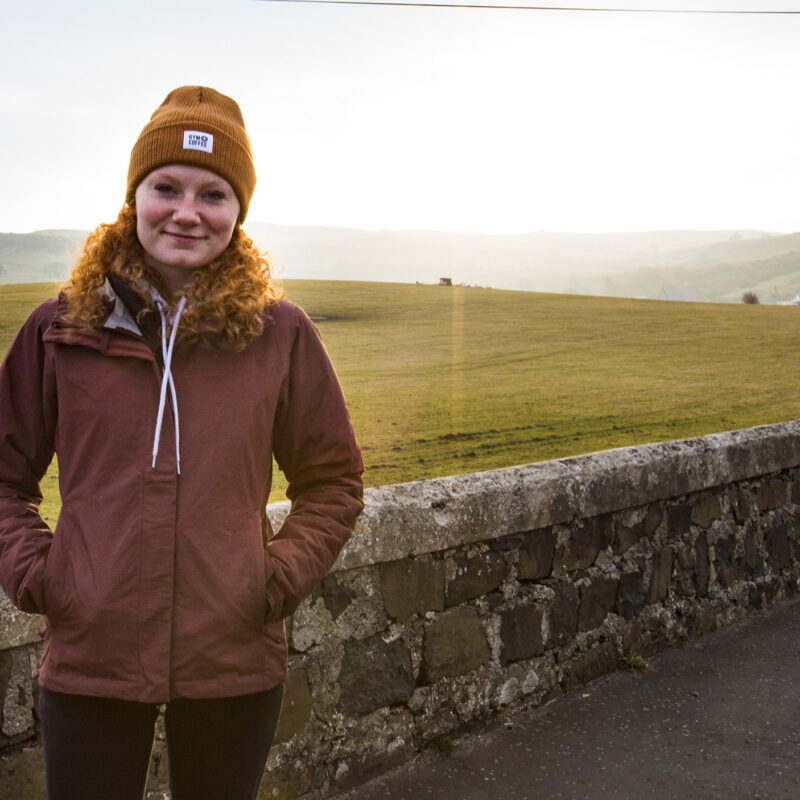 female student stands next to a rock wall, field and white houses in the background at sunrise