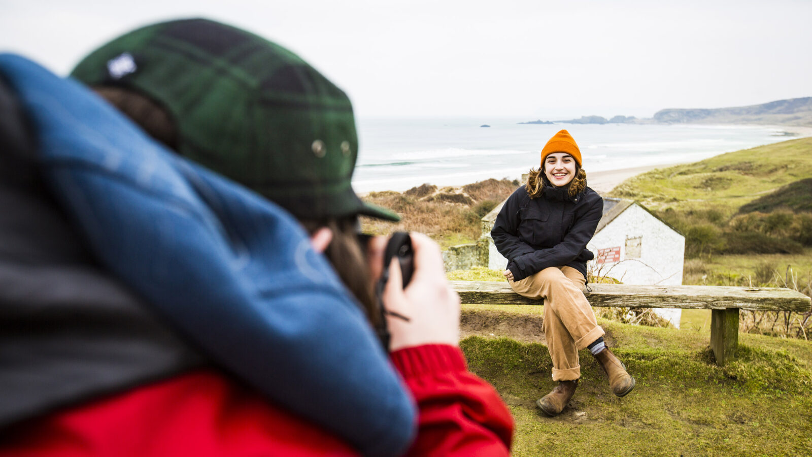one students takes a photo of another student as she is seated on a bench, ocean in the background