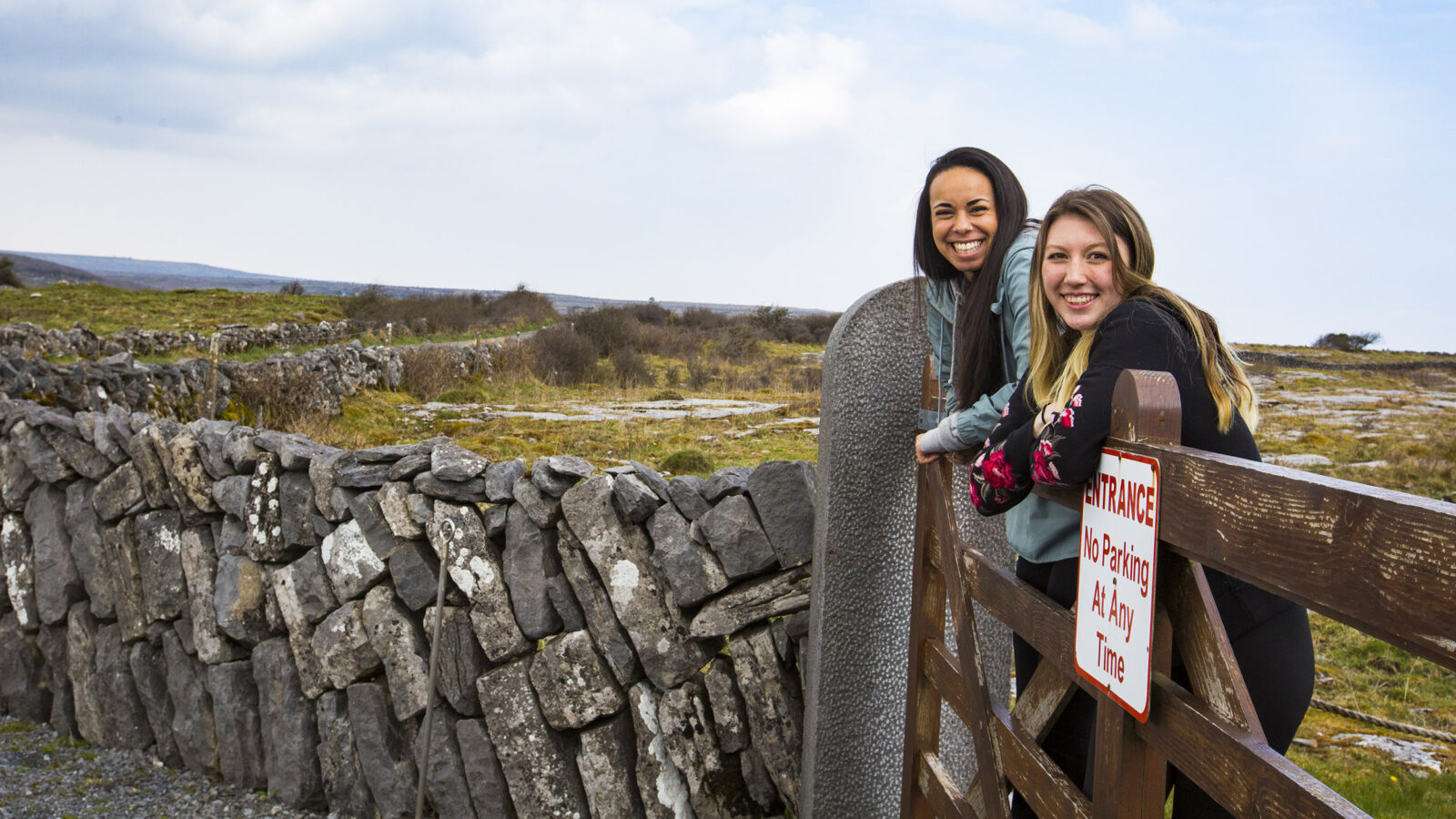 two students stand on a fence, a rock wall encloses a field