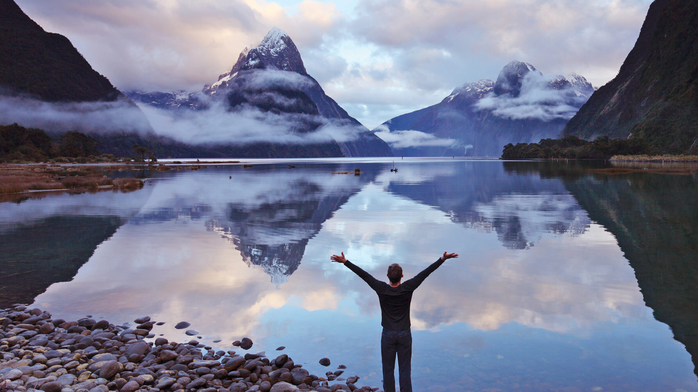 man with arms extended in the air looks at majestic mountains and lake