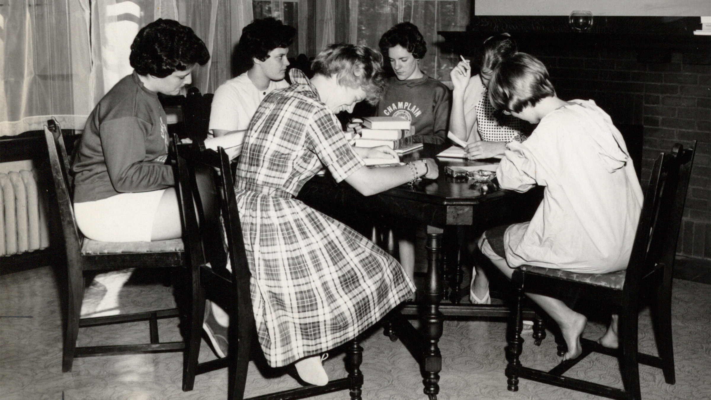 Black and white photograph depicting a group of Champlain College students studying in the common room of Jensen Hall. Six female students are seated at a table, bent over their textbooks and notes.