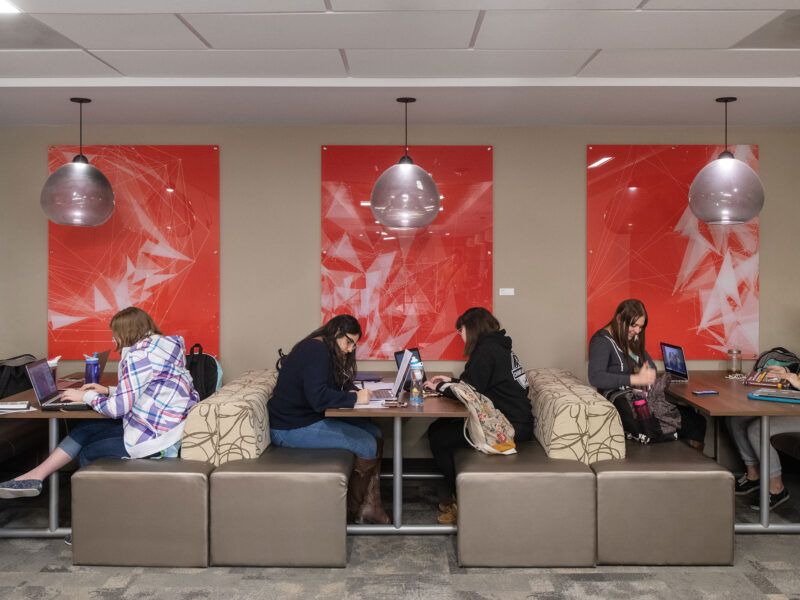 a wide angle of five students studying on their laptops sitting on bronze cushion seats in EATS Lounge on campus with big red posters in the background