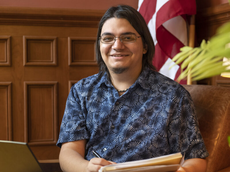 a student smiling at the camera while working in an office on a notebook and laptop