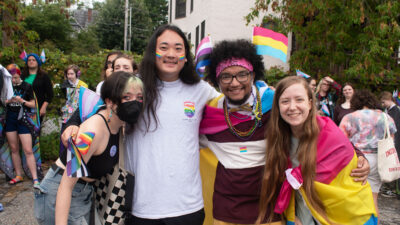 four students smile with their arms wrapped around each other during the burlington pride parade. two student wear colorful flags as capes and one student has rainbows painted on their cheeks