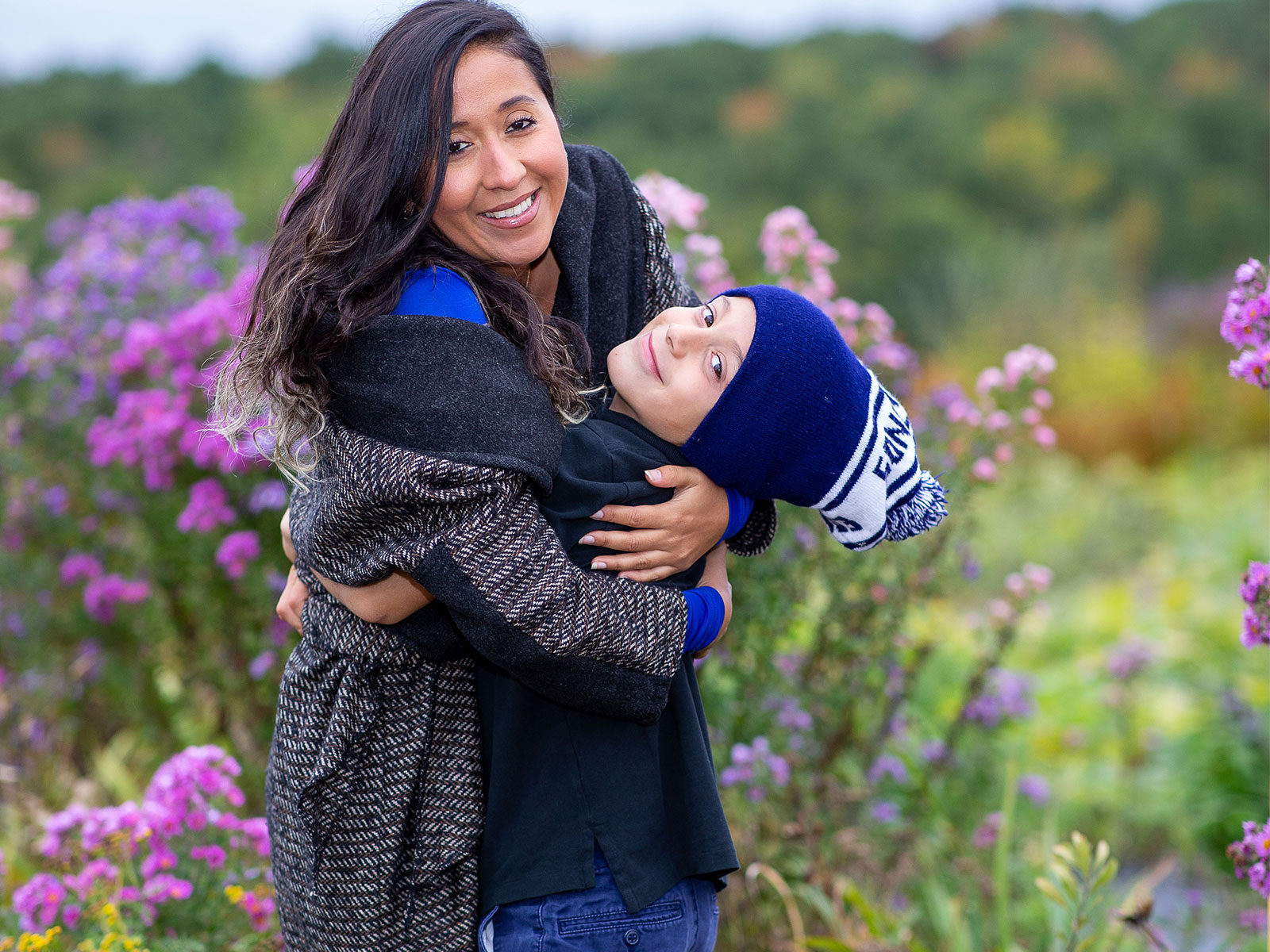 a mom hugs her child as they both smile in a green field with purple flowers